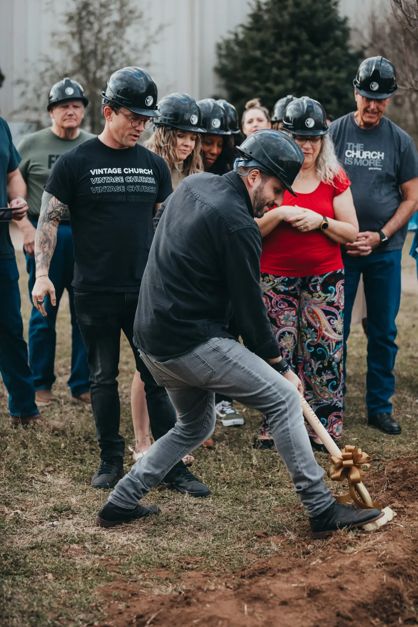 A man turns the first shovel of dirt as church members in hard hats look on at the groundbreaking