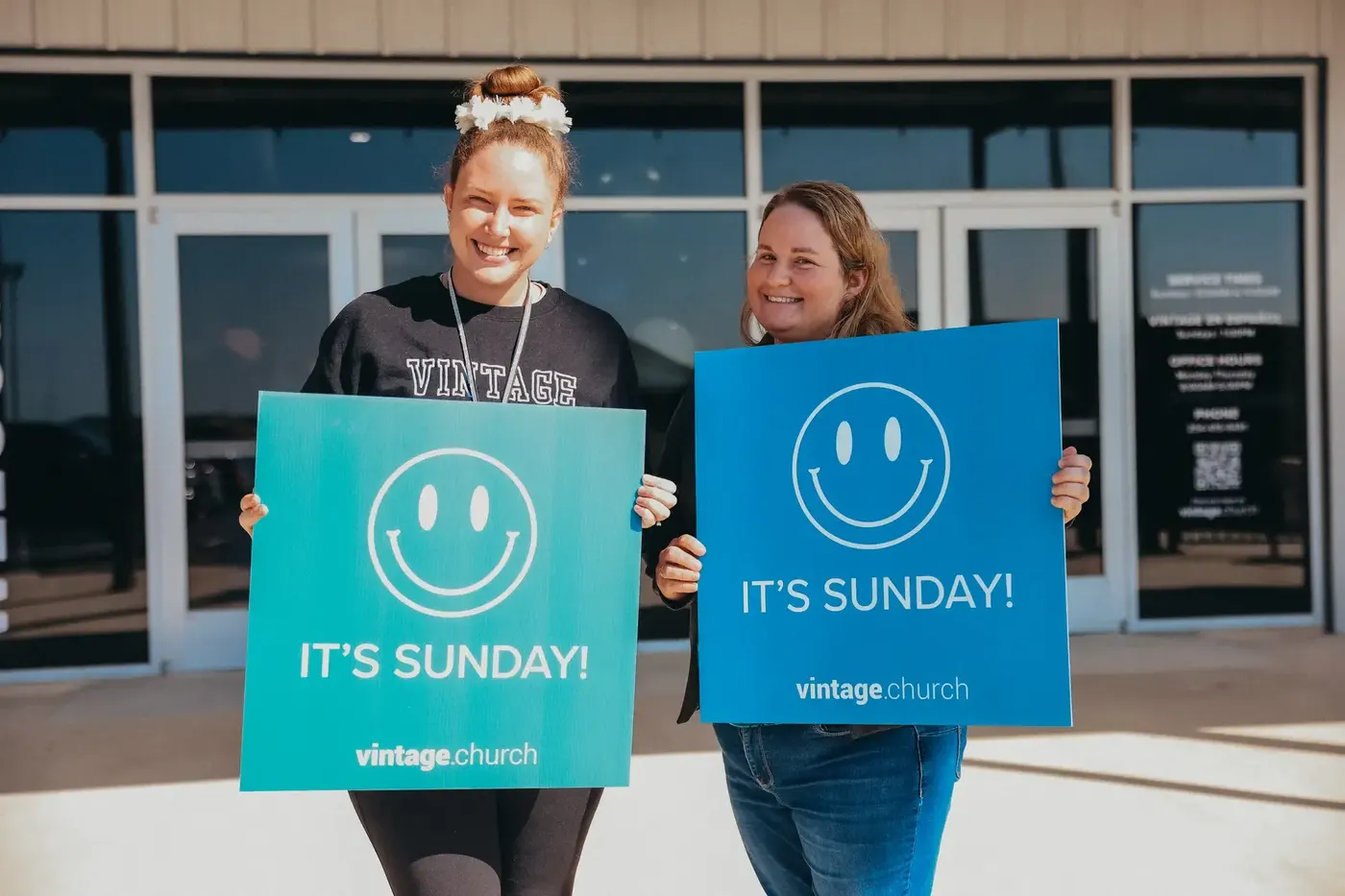 Two greeters holding It's Sunday signs outside the building