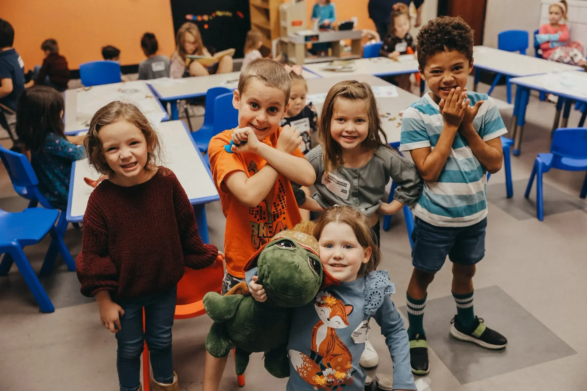 Smiling kids crowd together in a classroom, one hugging a stuffed green dinosaur