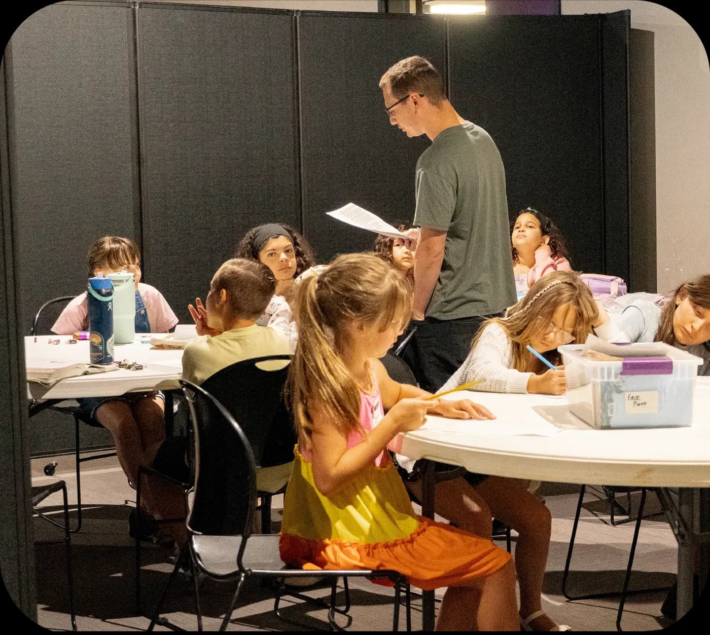 Kids work at round tables in an open room divided by portable black partitions
