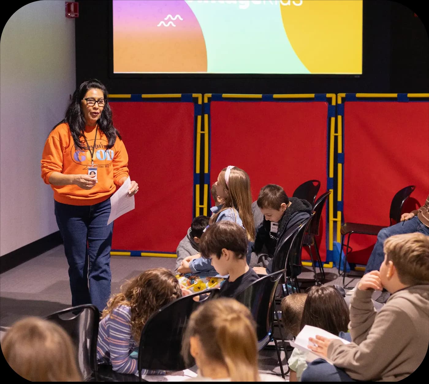 A teacher in an orange sweatshirt talks to kids seated on the floor of the auditorium in front of red padded panels