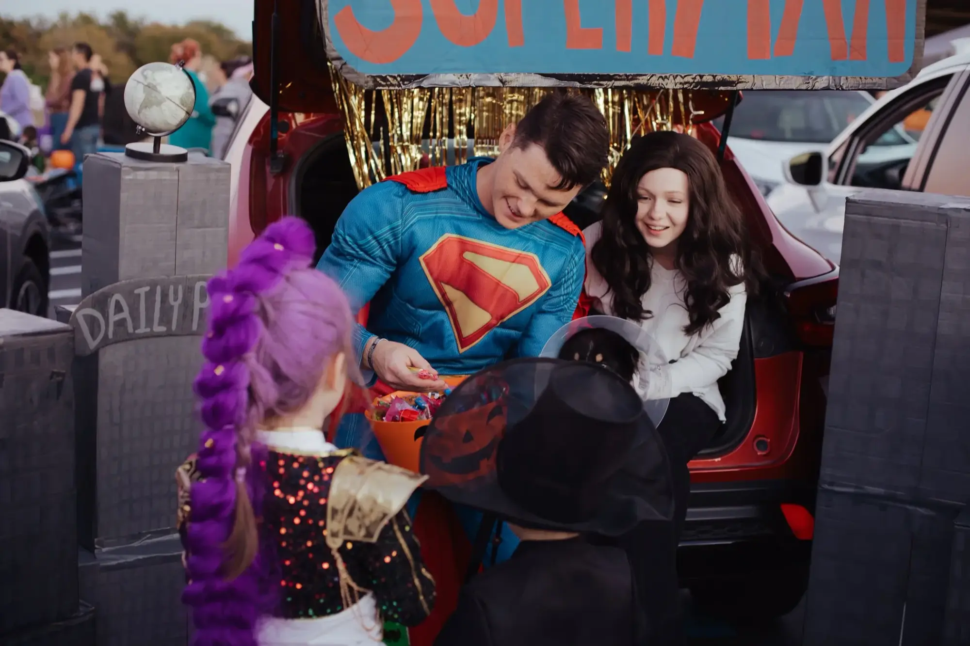 A volunteer dressed as Superman hands candy from a decorated trunk to children in costume