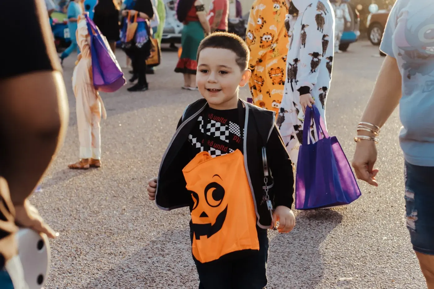 A smiling boy carries a jack-o'-lantern bag through the trunk-or-treat line