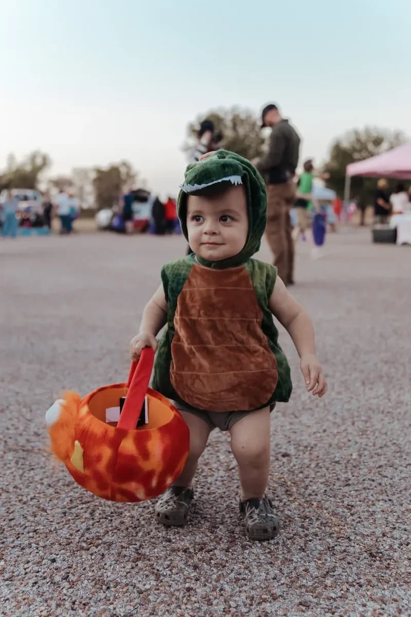 A toddler in a turtle costume walks across the lot carrying a pumpkin bucket