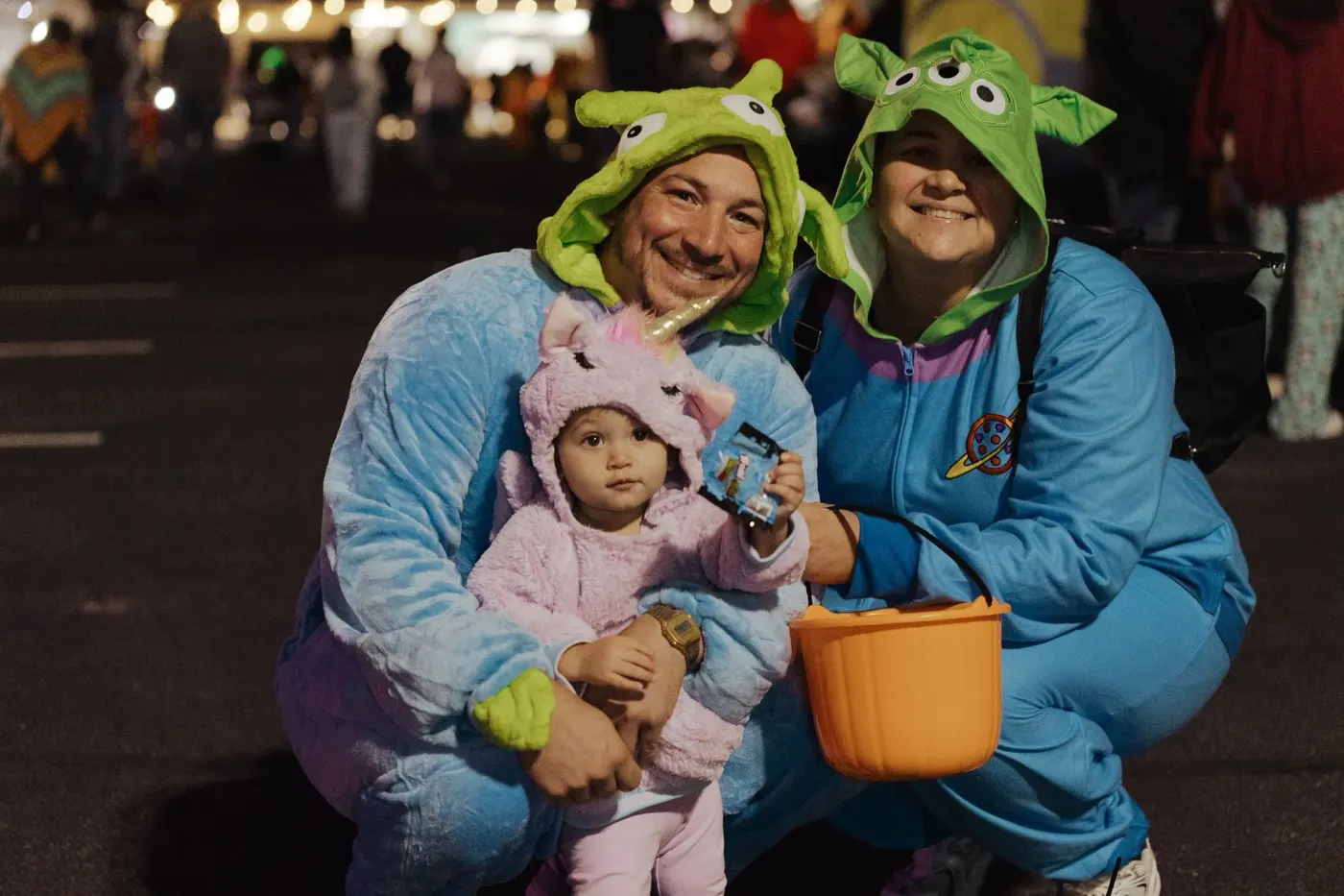 Parents in green alien costumes hold their baby, dressed as a pink unicorn