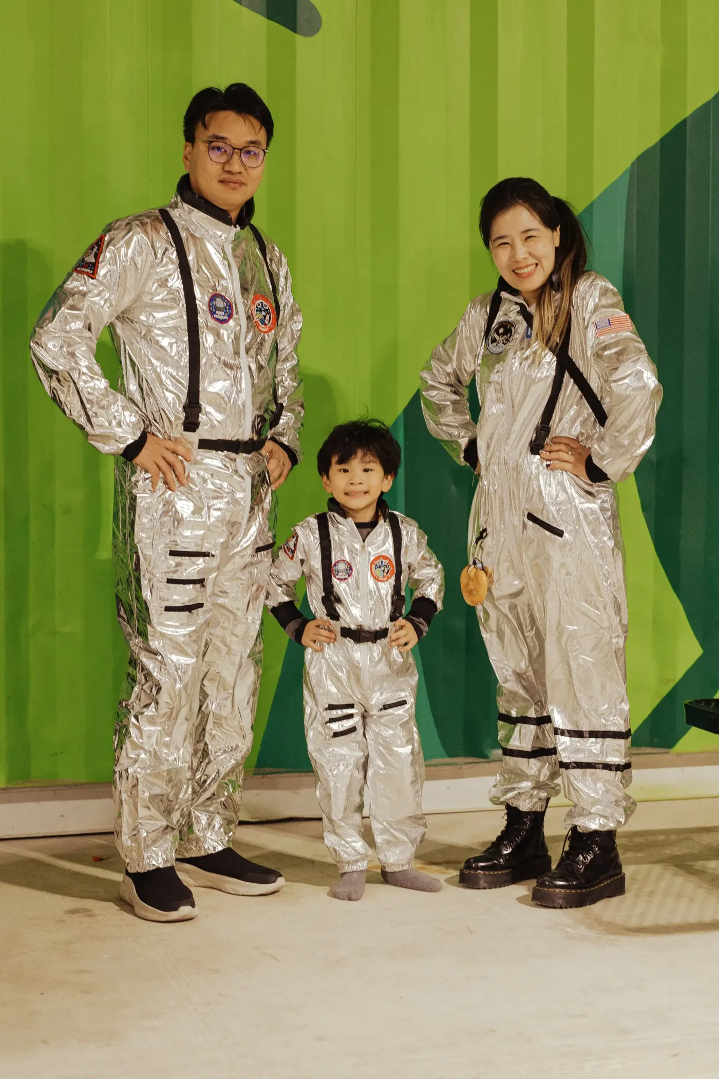 A family of three in matching silver astronaut suits poses against a green backdrop