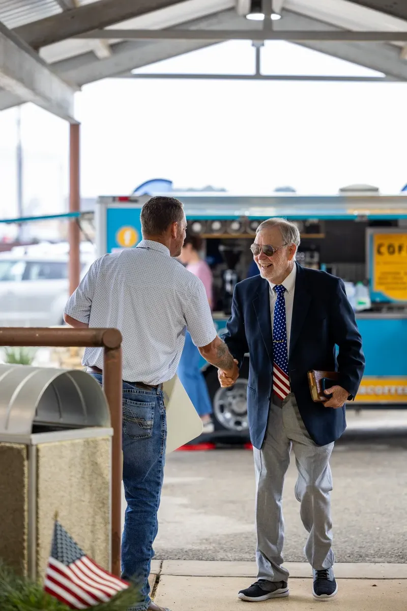 A greeter shakes hands with a smiling older man carrying a leather book outside, a coffee trailer behind them
